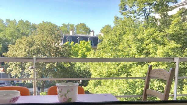 A balcony with a table and chairs, overlooking a green area with trees and buildings.