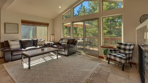 Uma sala de estar espaçosa com um sofá grande, uma mesa de centro e uma lareira. Há uma varanda com vista para árvores e um espaço de pátio com mobiliário exterior.