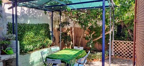 A covered patio with a table and chairs, surrounded by lush greenery and a wooden fence.