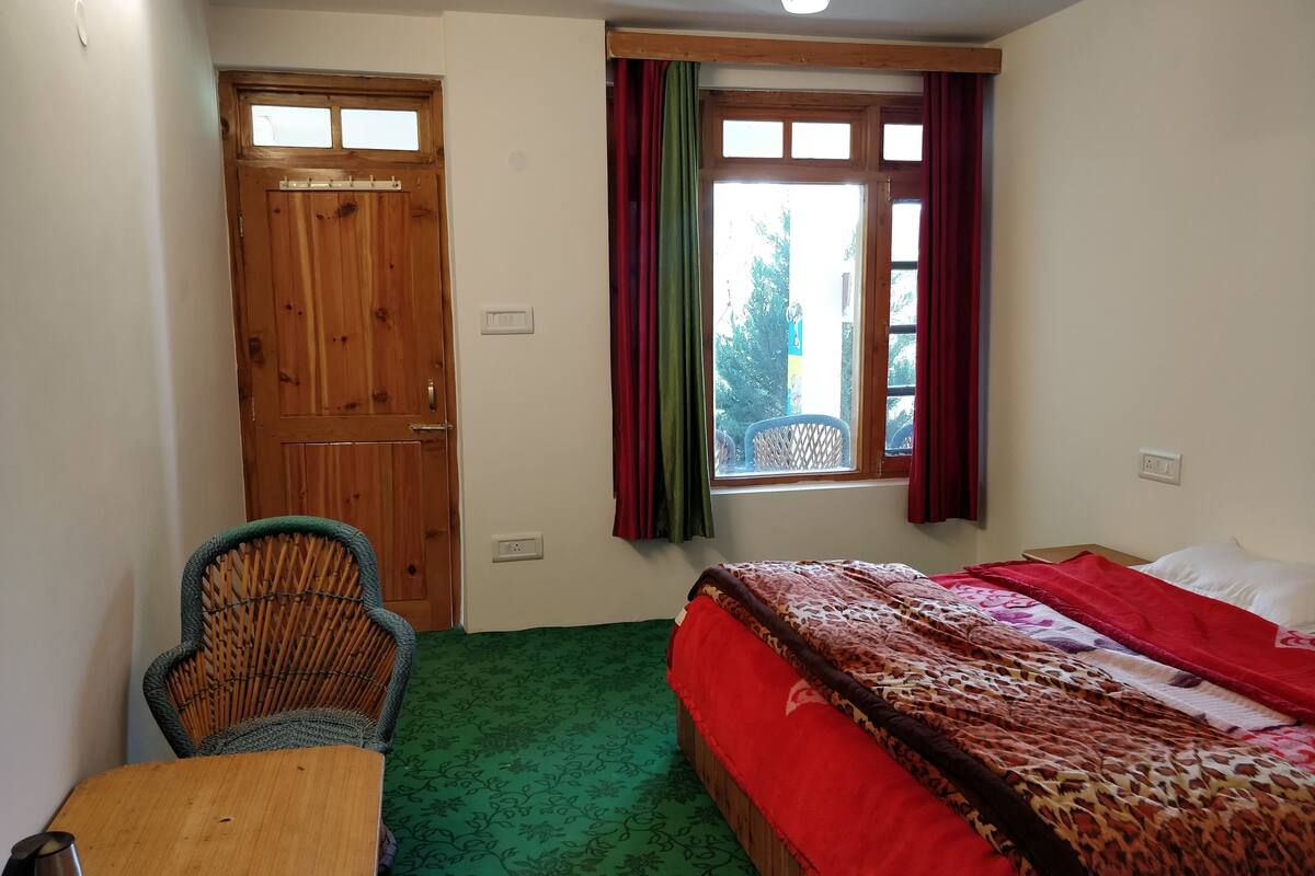 Una habitación con puerta de madera, ventana con cortinas rojas, cama con colcha estampada en rojo y marrón, alfombra de motivos verdes y una silla de mimbre.