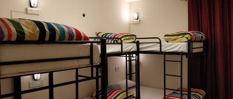 A dormitory room with bunk beds, each with a colorful striped blanket. There are wall-mounted lights and a power outlet on the wall.