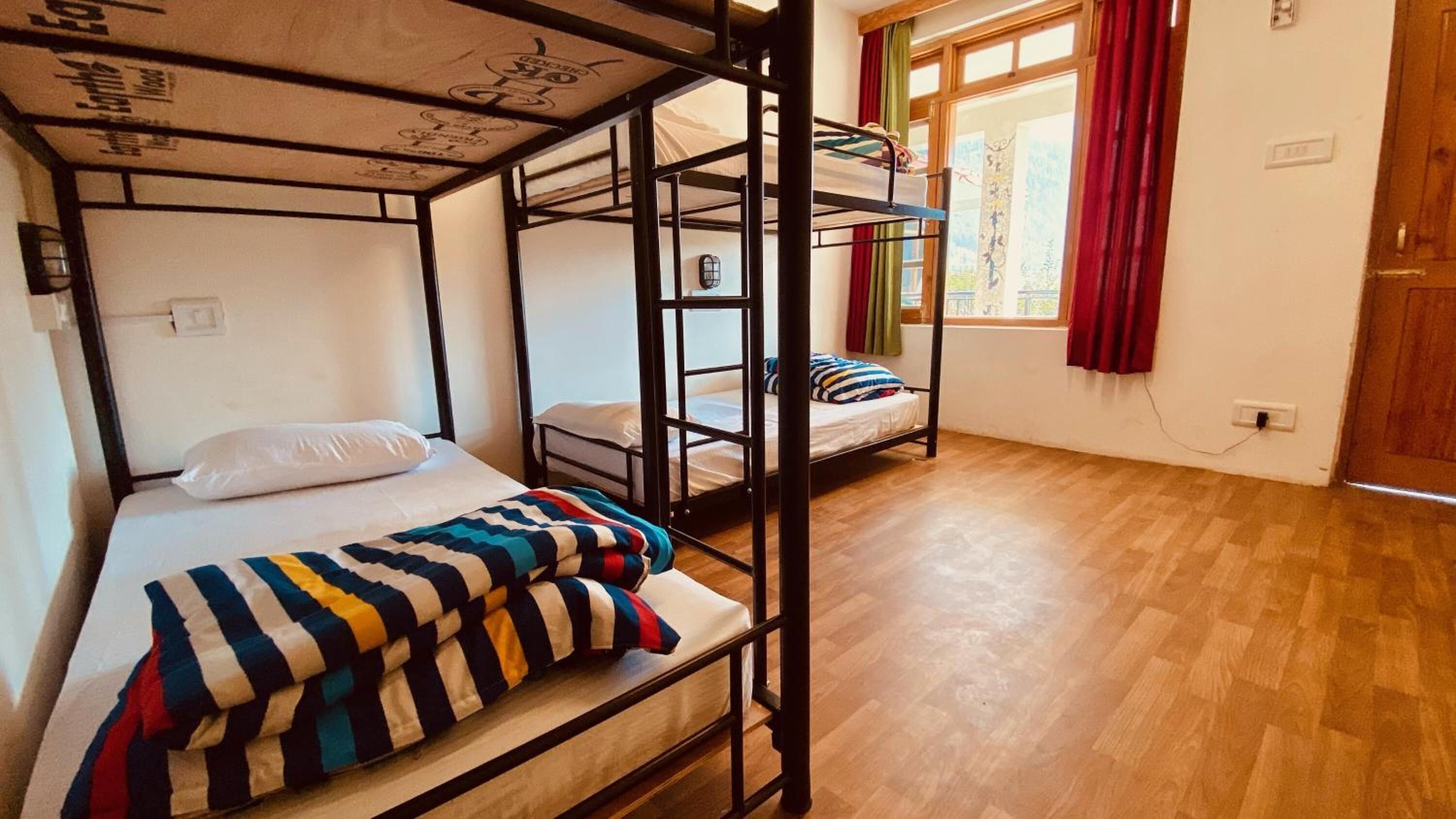 A dormitory room with bunk beds, wooden flooring, a window with red curtains, and a wall socket.