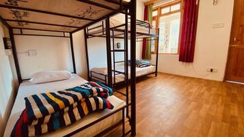 A dormitory room with bunk beds, wooden flooring, a window with red curtains, and a wall socket.