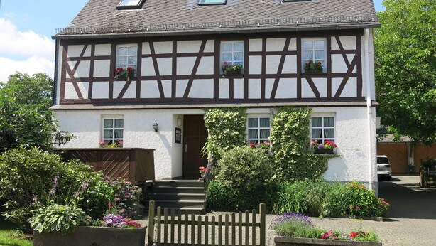 A traditional half-timbered house with a steeply pitched roof, dormer windows, and a front entrance with steps. - Cozy 2 pers. Vacation apartment "Sunflower" (Irmenach)