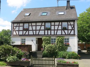 Une maison traditionnelle à colombages, dotée d’un toit à forte pente, de lucarnes et d’une entrée surélevée par quelques marches.