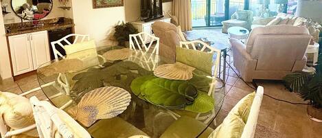 Uma sala de jantar com mesa de vidro, rodeada por cadeiras brancas, um candelabro e um ventilador de teto.