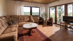 A living room with a sectional sofa, a coffee table, and a TV on a wooden stand. There are two chairs, a potted plant, and a view of the outdoors through large windows.