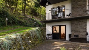 Une maison à deux étages avec une façade en pierre, un balcon doté de sièges d’extérieur et un mur de soutènement en pierre.