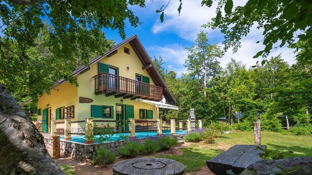 A two-story house with a balcony, a swimming pool, and a garden with a wooden table and a tree stump.