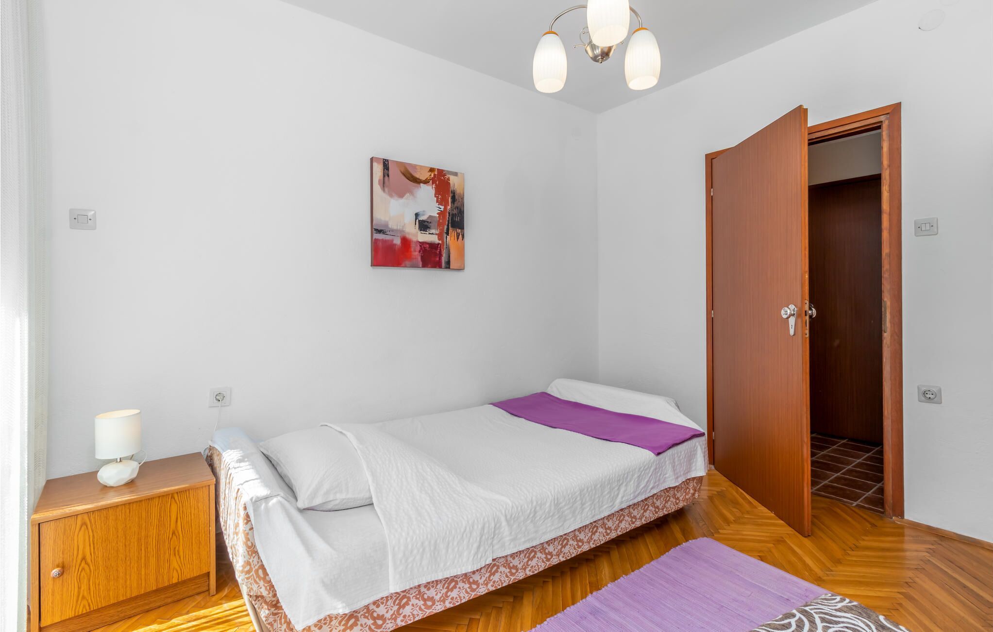 Un dormitorio pequeño y ordenado con cama, mesita de noche con lámpara, armario de madera y una puerta que da a otra habitación.