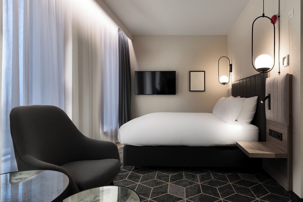 Een hotelkamer met een bed, een stoel, een klein tafeltje, een televisie en een wandlamp.