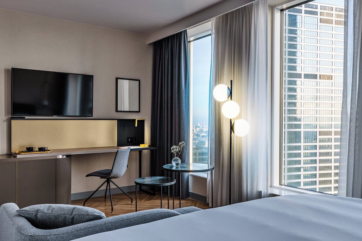 Een moderne hotelkamer met een bed, een bureau, een stoel, een kleine tafel, een televisie en een groot raam met gordijnen.