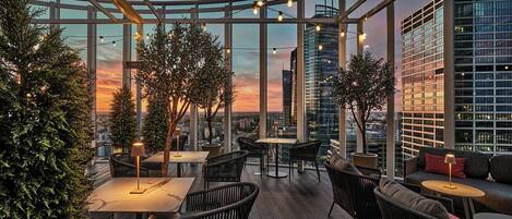 Una terraza en la azotea con mesas y sillas, plantas en macetas y vista panorámica de la ciudad.