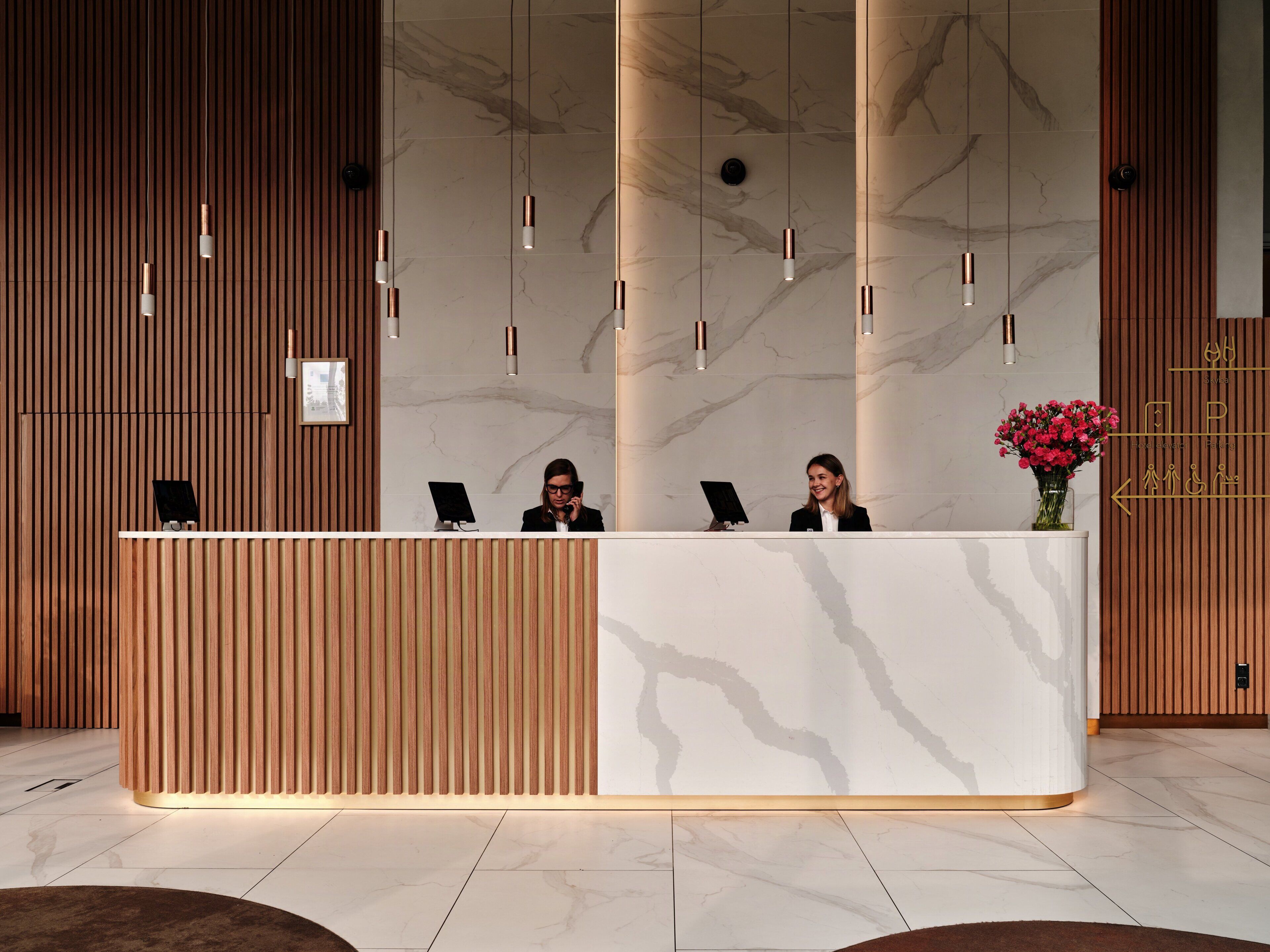 A reception desk with a marble wall and wooden paneling, two receptionists, and a flower vase.