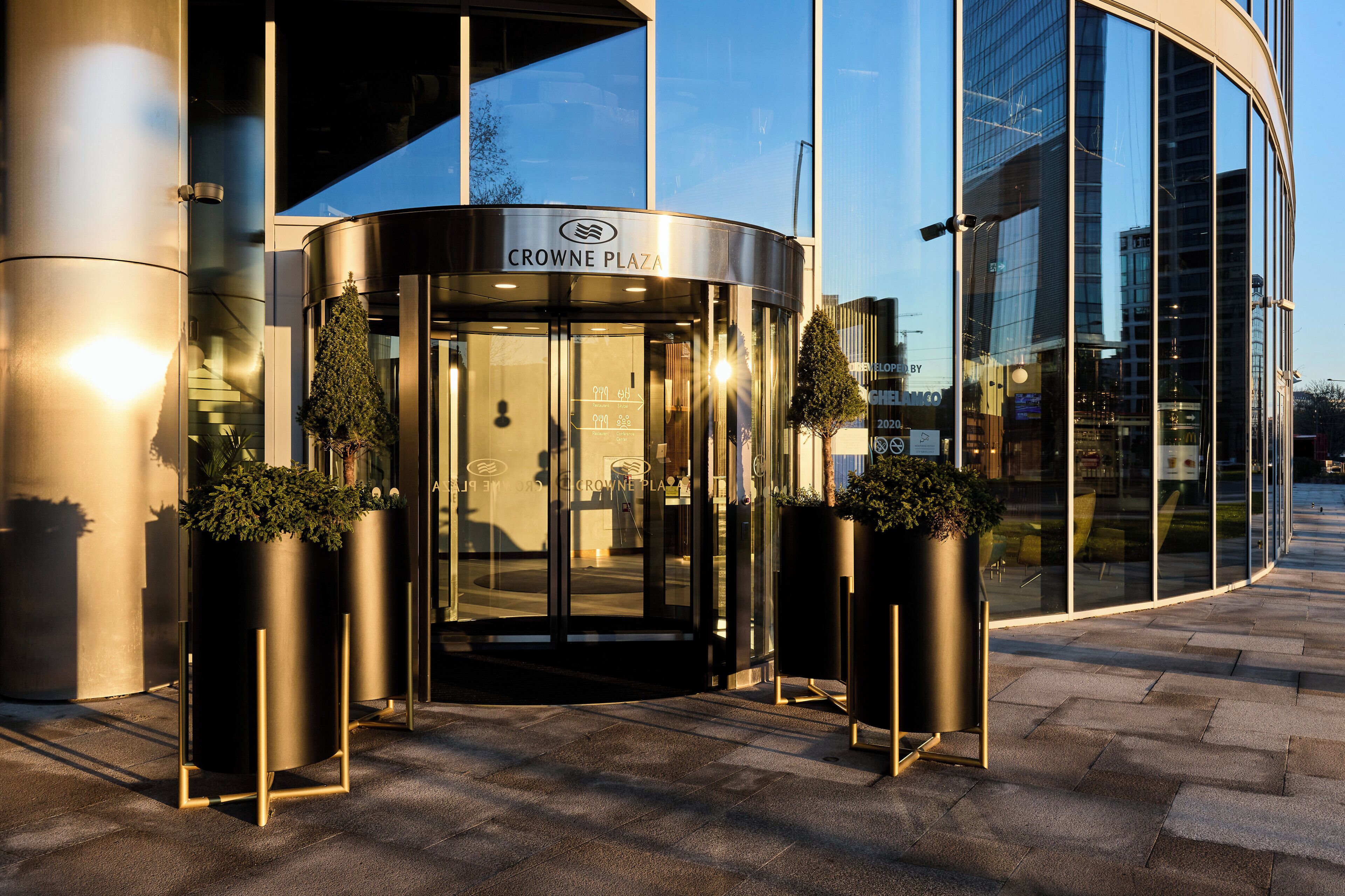 Fachada moderna de edificio con puerta giratoria, paredes de cristal y plantas en macetas.