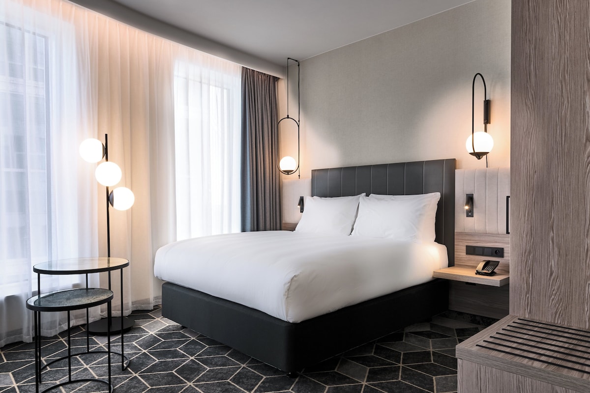 Een hotelkamer met een groot bed, een nachtkastje, een lamp en uitzicht op de stad.