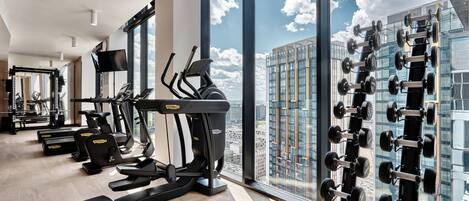 A gym with treadmills, ellipticals, and weight machines, large windows with a city view.