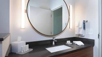 Une salle de bain moderne avec un miroir rond, un lavabo blanc et un plan de toilette foncé.