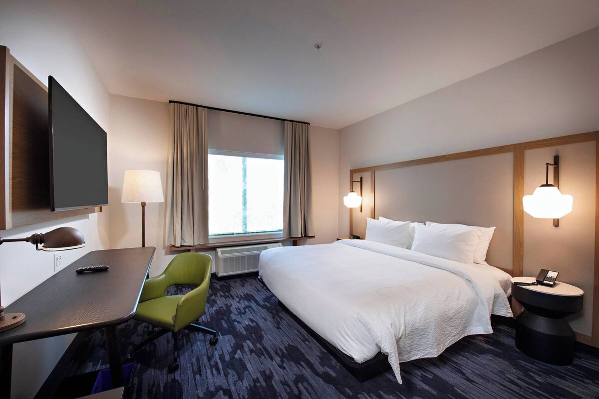 Une chambre d’hôtel avec un grand lit, un bureau avec une chaise, une télévision et une fenêtre avec des rideaux.