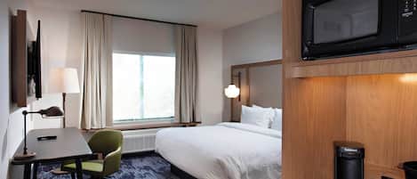 A hotel room with a bed, a desk, a chair, a lamp, a window with curtains, a microwave, and a coffee maker.