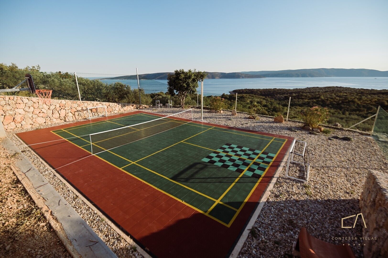 Un court de tennis avec un filet, entouré d’un mur de pierre, et offrant une vue sur un lac et des collines.