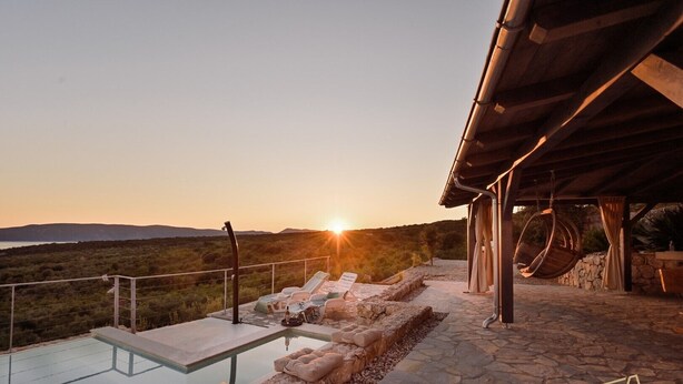 Una piscina infinita con sillones de descanso, un camino de piedra y vistas al atardecer sobre un paisaje montañoso.