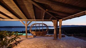 Un patio de piedra con una silla mecedora bajo una pérgola de madera, con vistas a un cuerpo de agua y colinas.