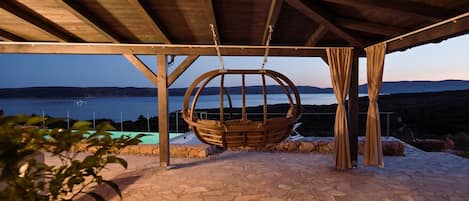 A stone patio with a swing chair under a wooden pergola, overlooking a body of water and hills.