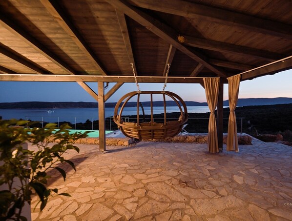 Eine Steinterrasse mit Schaukelstuhl unter einer Holzpergola, mit Blick auf ein Gewässer und Hügel.