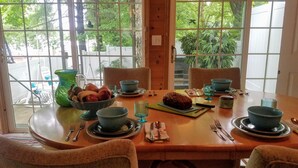 Een eettafel gedekt met kommen, borden, bestek en een brood, met uitzicht op groen buiten.
