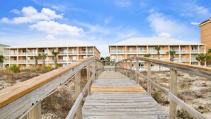 A wooden boardwalk leading to a beachfront hotel with multiple balconies and palm trees.