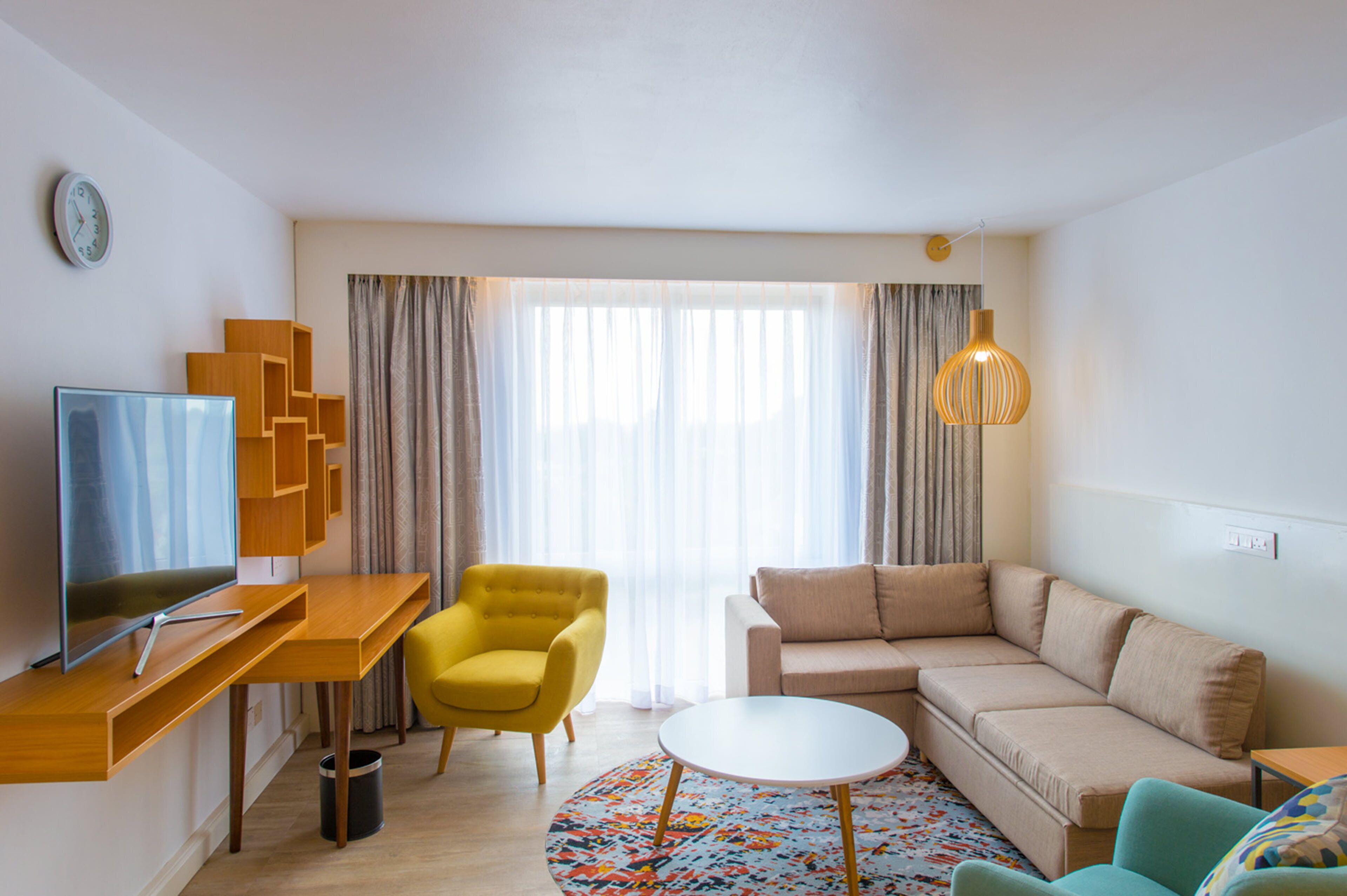 Una sala moderna con televisor, un sillón amarillo, un sofá color crema, una mesa redonda y una alfombra estampada.