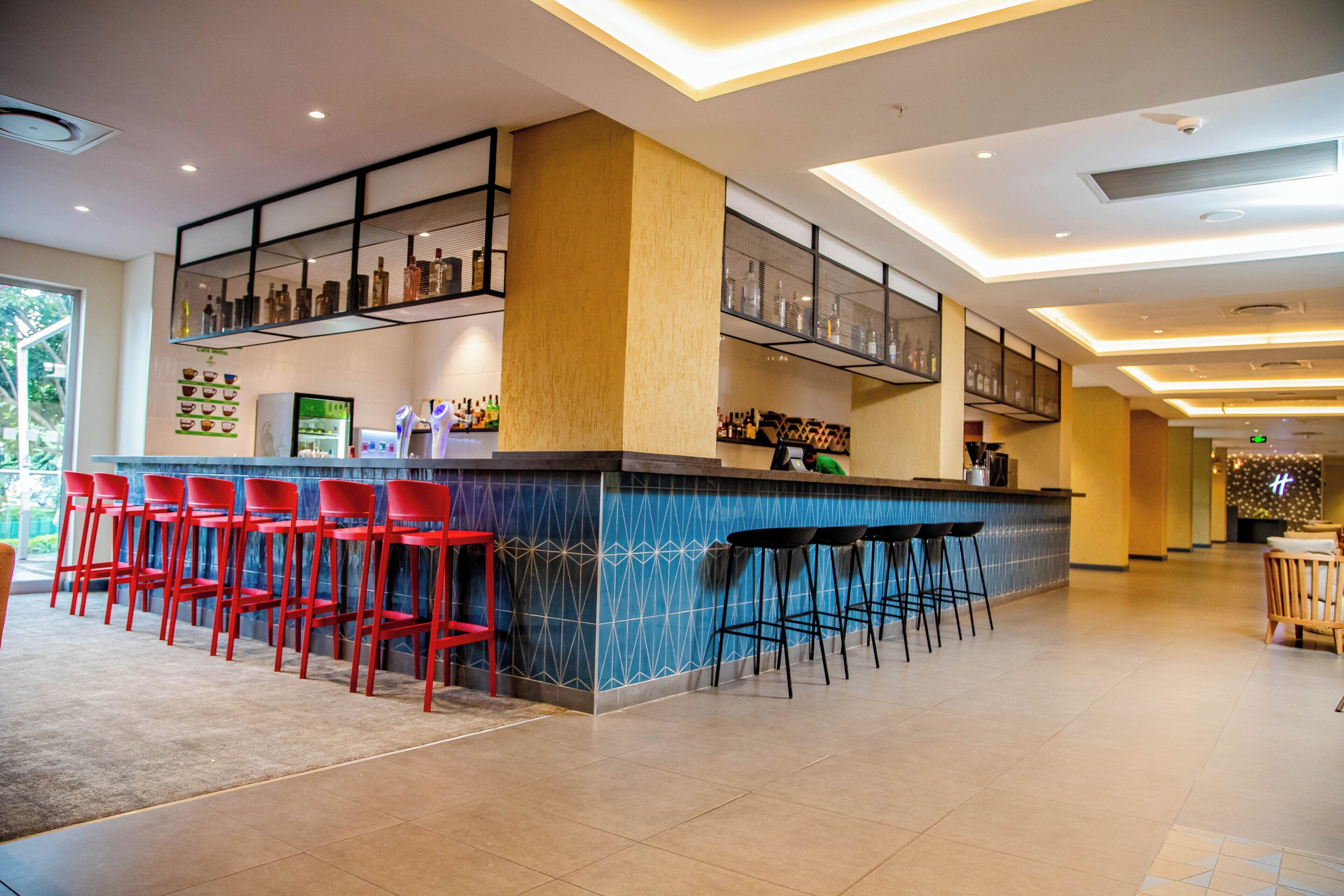 Zona de bar con taburetes negros y rojos, una barra con protector contra salpicaduras azul estampado, y estantes con botellas.