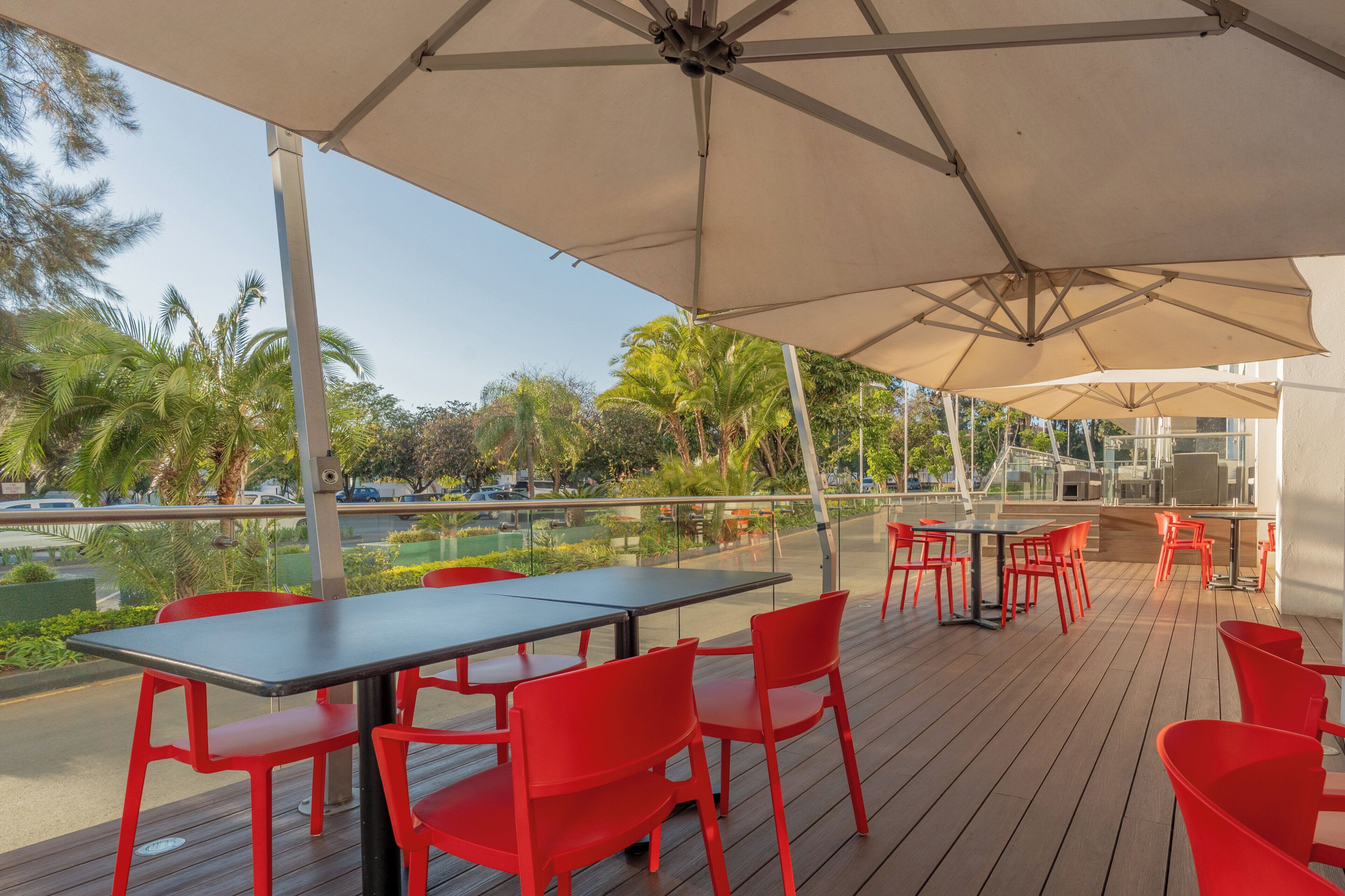 An outdoor patio with a table and chairs, a large umbrella, and a view of palm trees and a parking lot.