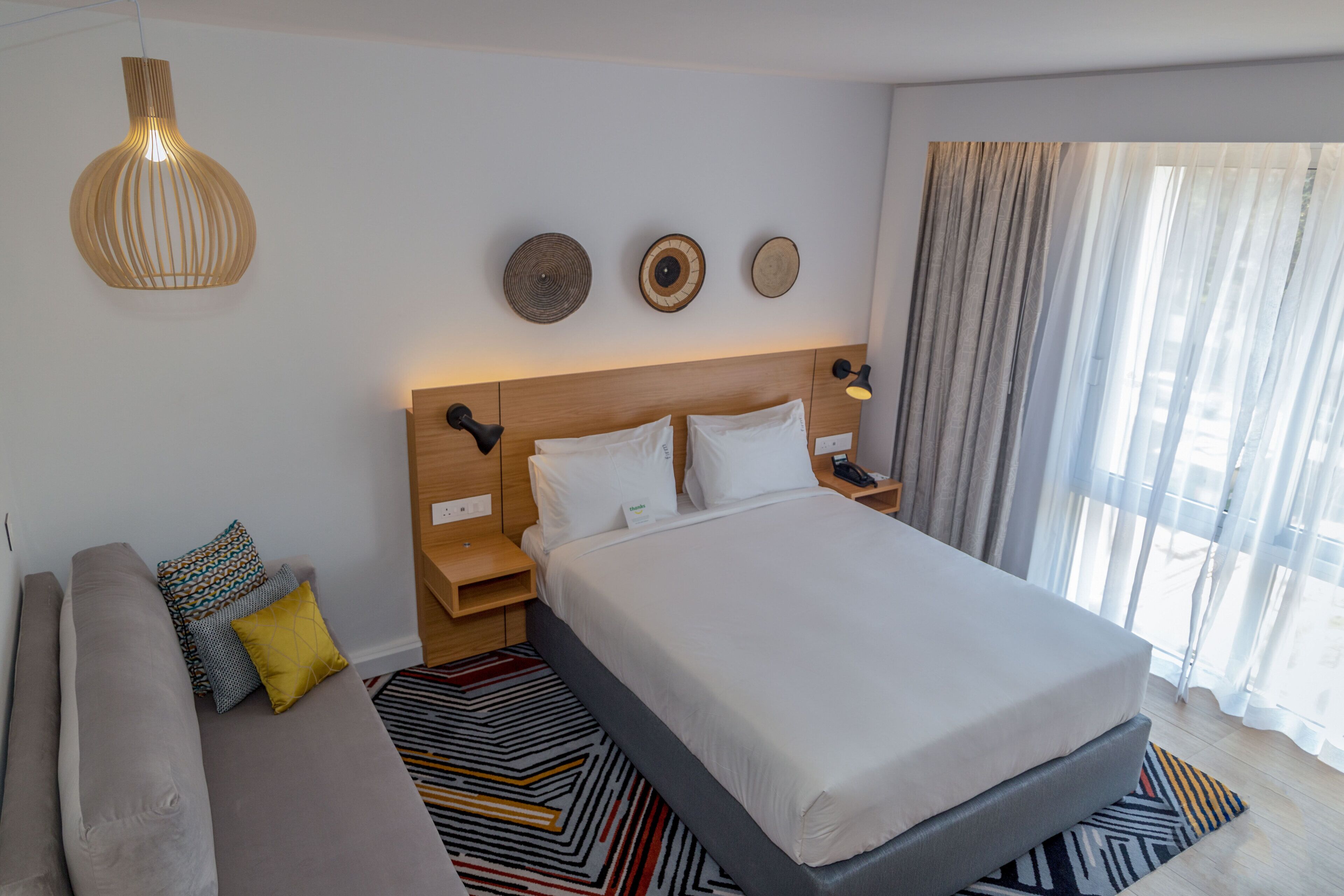 A hotel room with a large bed, a sofa, a bedside table, a lamp, and a window with curtains.