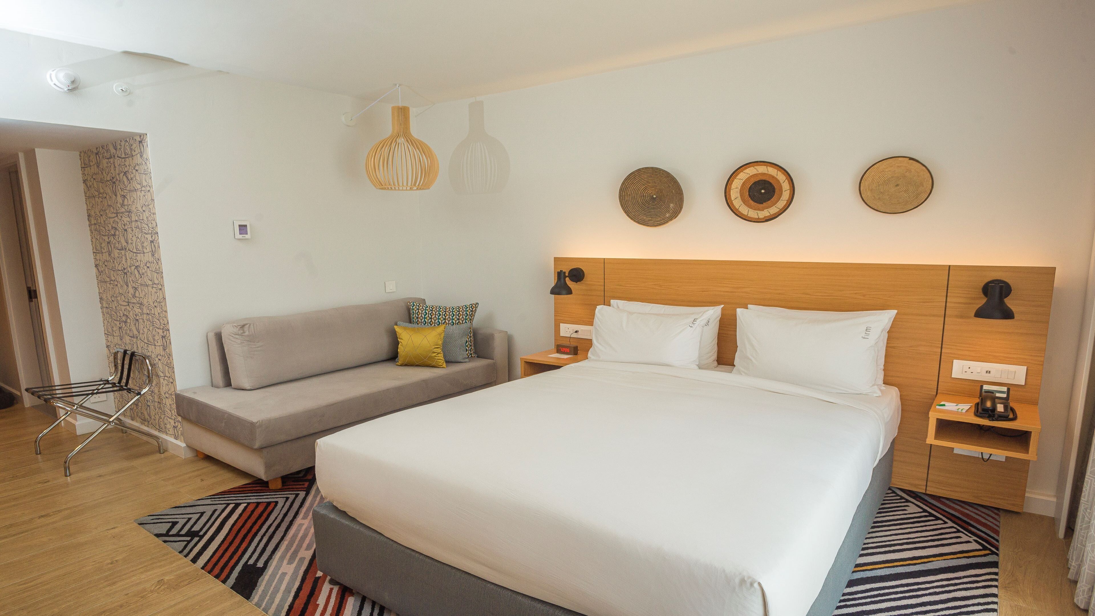 A hotel room with a large bed, a sofa, a chair, and a bedside table with a lamp.