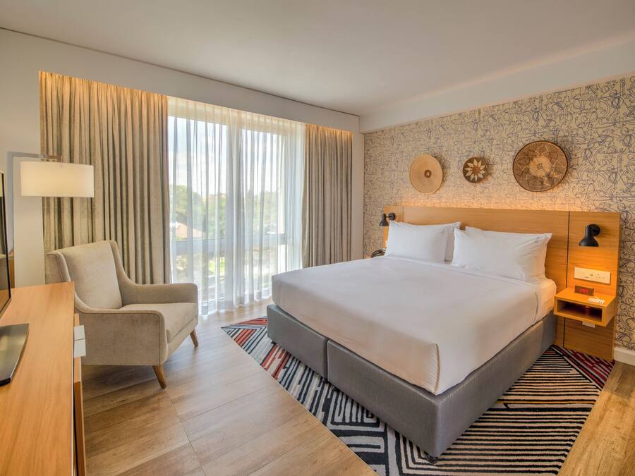 A modern hotel room with a large bed, a desk, a chair, and a view of the outdoors through large windows.
