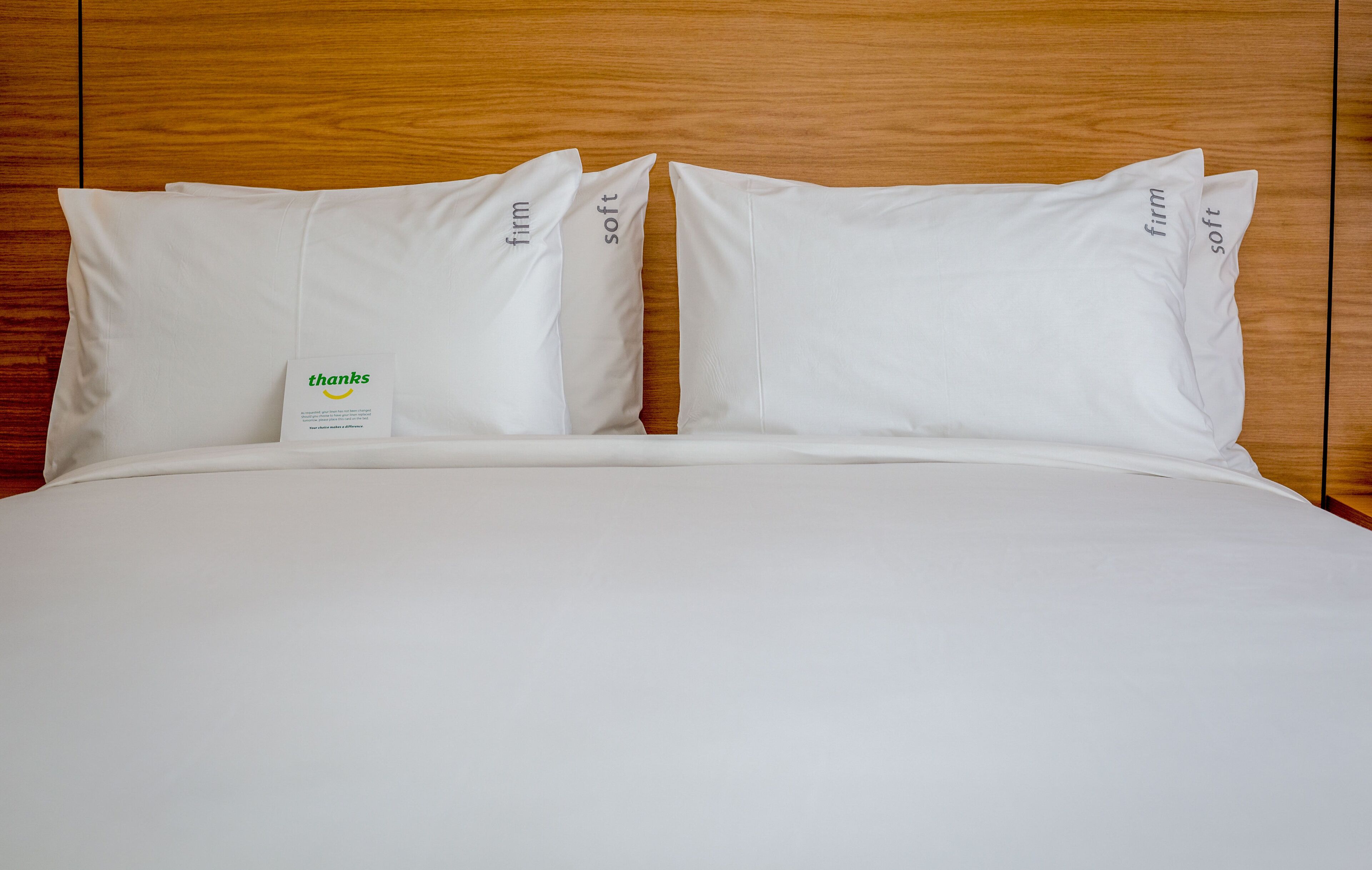 A neatly made bed with white bedding and pillows, a wooden headboard, and a small card with the word 'thanks' on it.