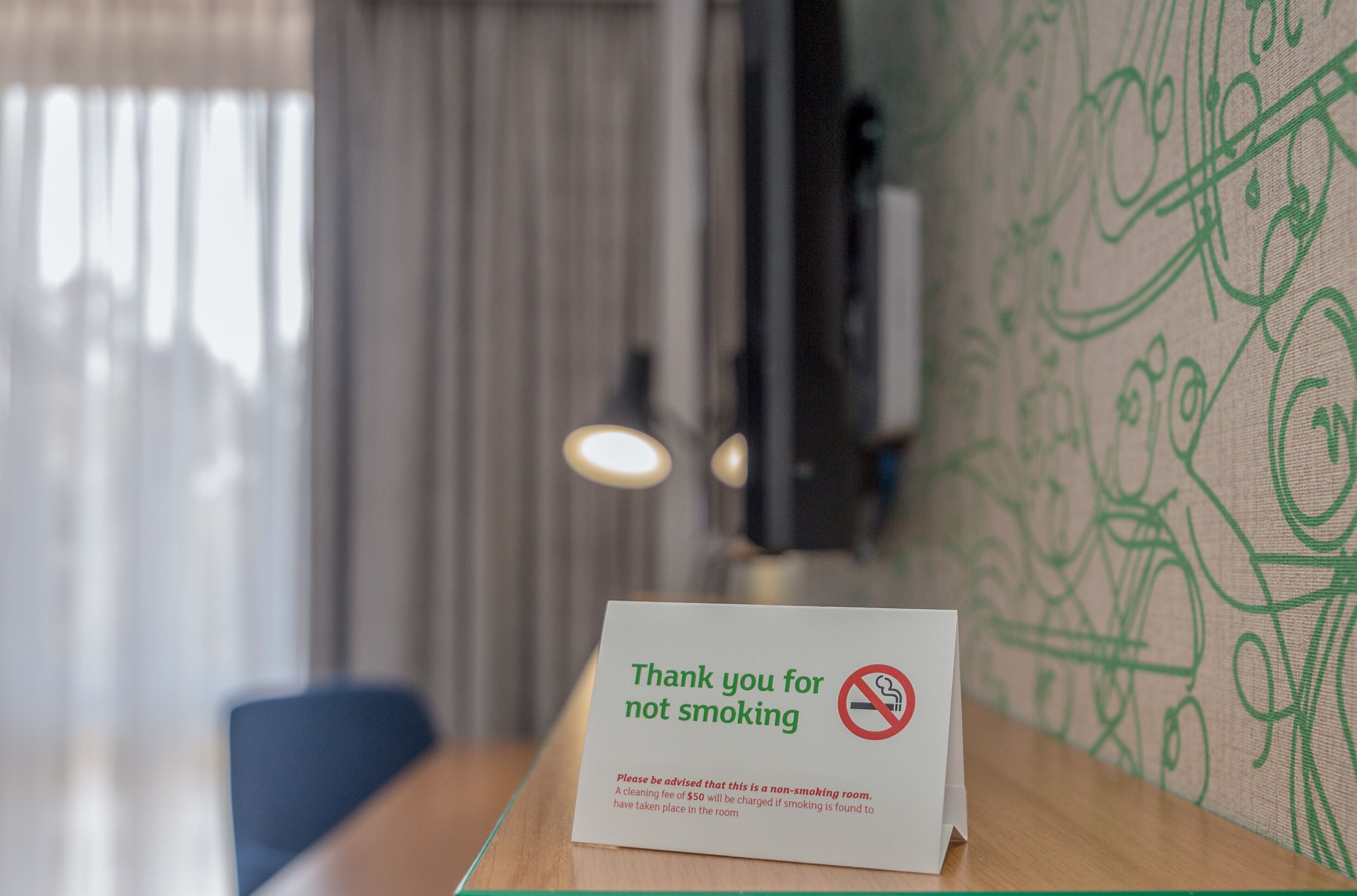 Señal de prohibido fumar sobre una mesa en una habitación con pared estampada y ventana con cortinas.