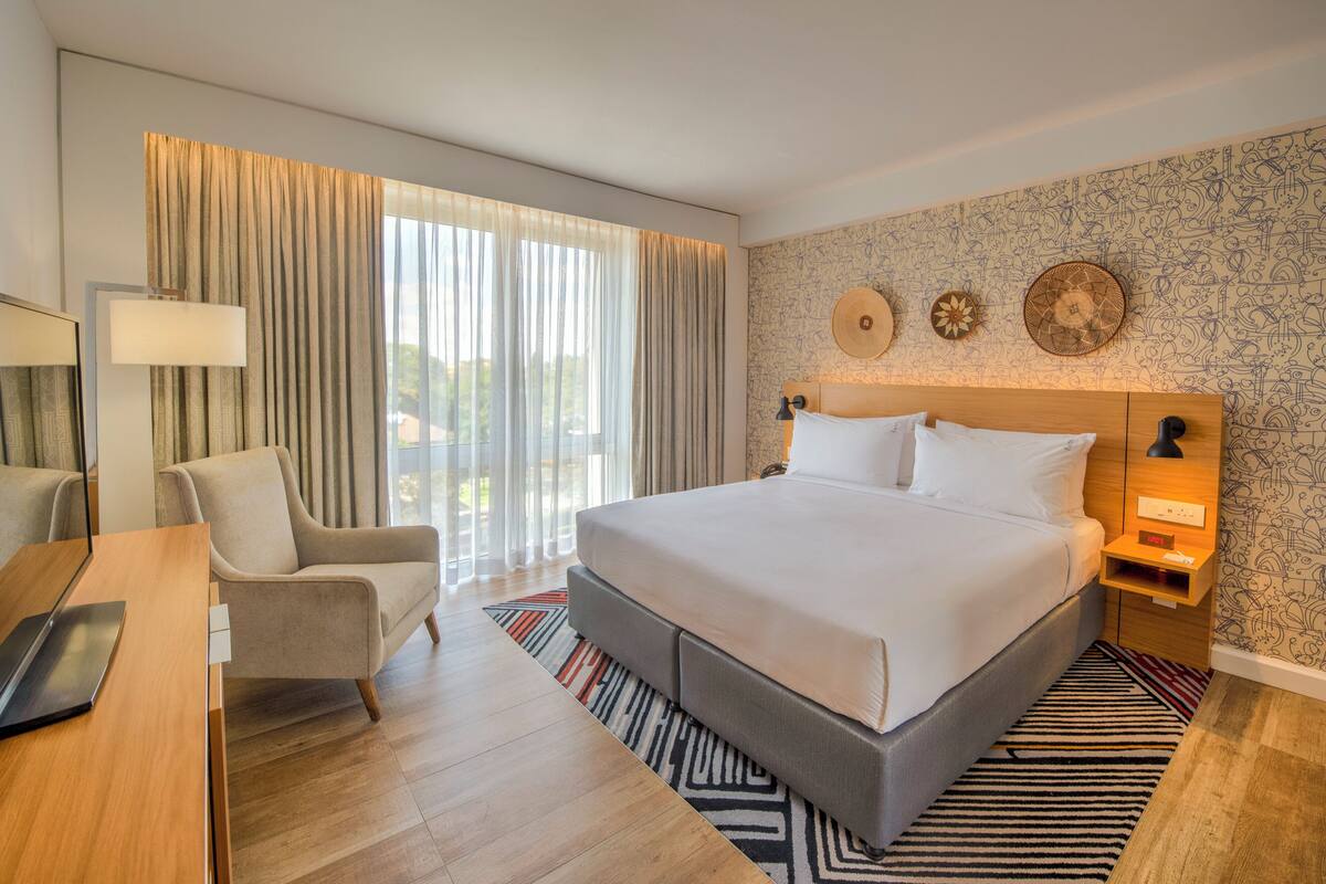Camera d'hotel moderna con un ampio letto, una scrivania, una sedia e vista all'esterno tramite ampie finestre.