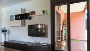 A modern living room with a flat-screen TV, built-in shelves, a dark entertainment unit, and a sliding glass door leading to an outdoor area with potted plants.