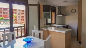 A modern kitchen with a central island, a dining area with a white table and chairs, and a view of a residential building through a glass door.