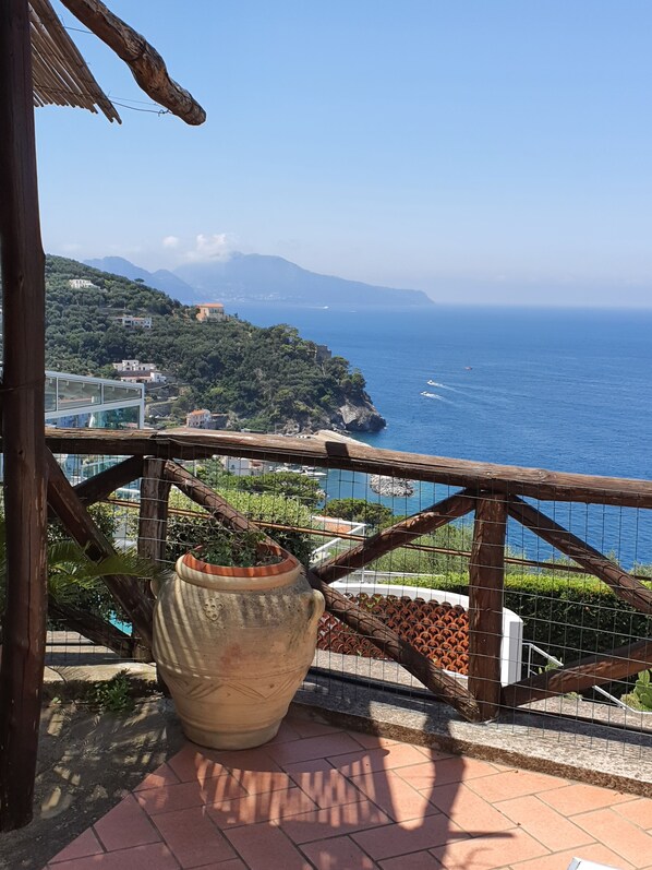 Un balcón con una maceta de terracota grande, barandilla de madera y vista al mar y edificios distantes.