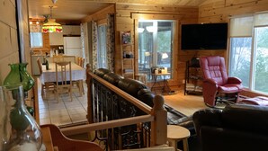 A cozy cabin interior with a dining area, a living room with a leather sofa, a red armchair, and a TV mounted on the wall.
