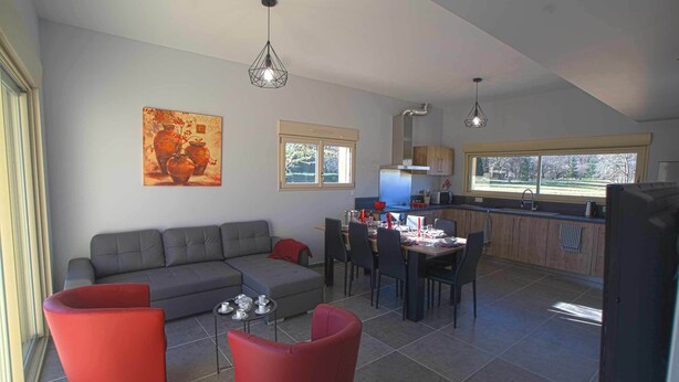 Ein modernes Wohnzimmer mit grauem Sofa, roten Sesseln und einem Essbereich mit Tisch und Stühlen. Im Hintergrund ist eine Küche mit einem Fenster und Blick auf Bäume zu sehen.