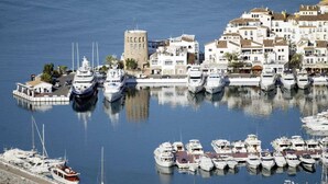 En småbåtshamn med flera yachter förtöjda, ett markant torn och vita byggnader med balkonger.