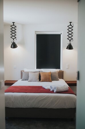 Una cama bien hecha con una colcha roja y varias almohadas, un ventanal grande con una persiana negra y dos lámparas colgantes negras a cada lado.