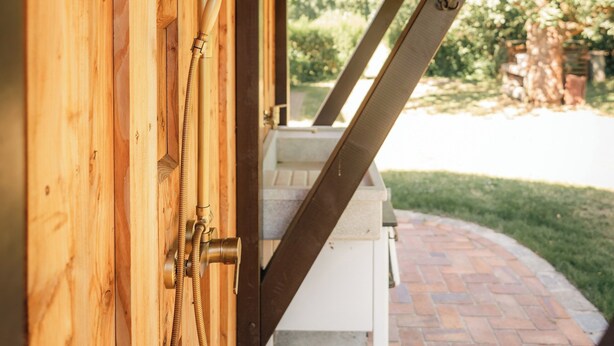 A wooden structure with a metal showerhead and hose, a white bench, and a brick pathway leading outside.
