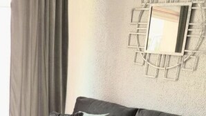 A grey sofa with multiple cushions, a glass coffee table, and a grey rug in a room with a textured wall and a geometric mirror. - Studio Center Cannes Terasse (Cannes)