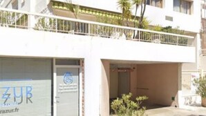 A white two-story building with a closed shutter on the ground floor, a balcony with railings, and a small garden area with plants. - Studio Center Cannes Terasse (Cannes)